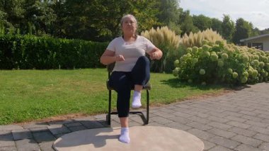 Aşırı kilolu bir kadının bahçede sandalyede otururken mekik çekişini ön plana çıkarmak karın, bacaklar ve kolları güçlendiriyor. Sağlıklı bir yaşam tarzı arayan yaşlılar için ev ödevi planı