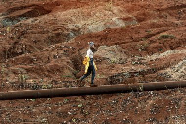 Sarı yağmurluklu genç bir kadın, kırmızı kil ocağının arka planında, bir kablo köprüsünde duruyor.