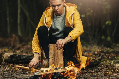 Sarı yağmurluklu erkek gezgin ormanda kamp ateşinin yanında oturuyor. Ormanda karanlık sonbahar havası. Ateşe odun hazırlıyor.