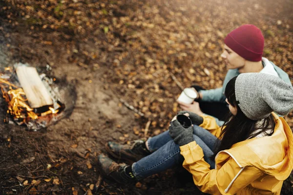 Young happy couple travellers with happy emotions drinking cup of hot drink next to camp fire in forest/ Dark autumn mood in the woods.