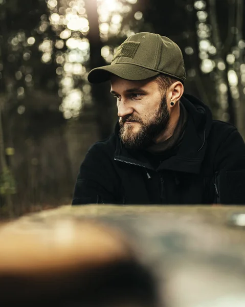 Bearded man with army green hat and black sweater outdoors .