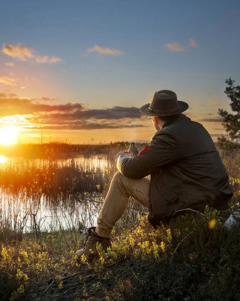 男性旅行者 戴着帽子坐在沼泽地里 夏天日落时喝热水瓶中的热饮 — 图库照片