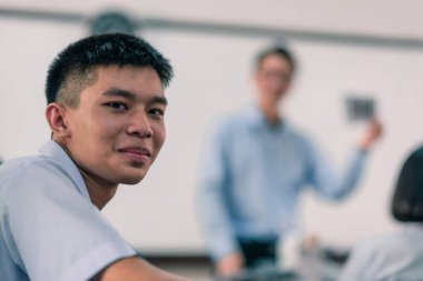 Beyaz üniformalı Asyalı bir erkek lise öğrencisi sınıfta öğretmenlik yaparken gülümseyen yüzünü geri çevirir..