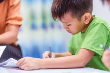 Asyalı ilkokul öğrencisi, sarı tişörtlü Asyalı bir bayan öğretmenle aynı sınıfta okuyor..