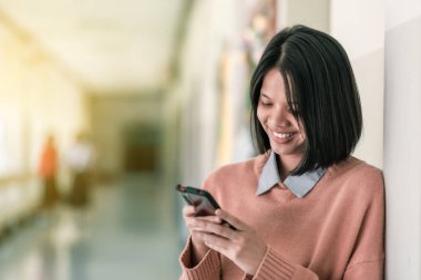 Gülümseyen Asyalı bir lise öğrencisi okuldaki akıllı telefonuyla sosyal medyanın tadını çıkarıyor..