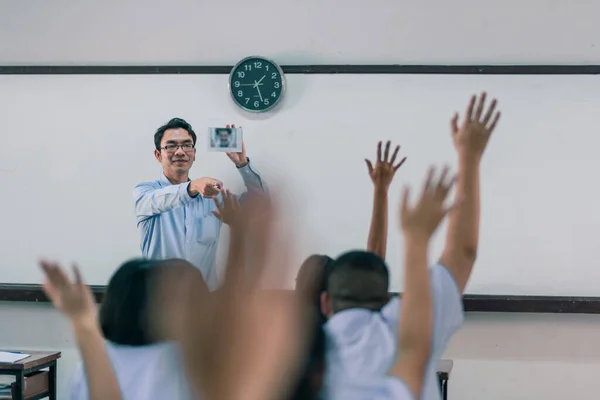 笑顔のアジア系男子高校生の先生が教室で制服を着た生徒たちに質問をし 生徒たちが手を挙げて答えを教えています — ストック写真