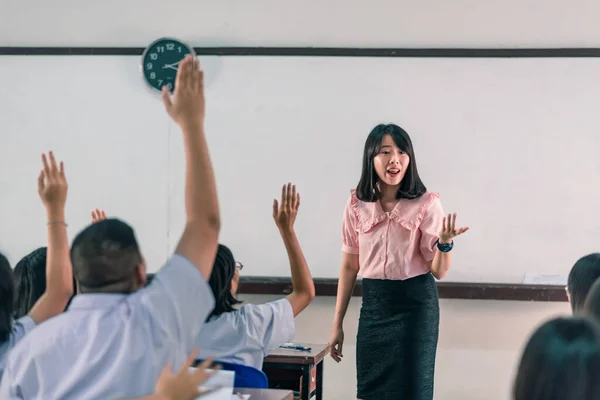 Gülümseyen Asyalı bir bayan lise öğretmeni sınıftaki beyaz üniformalı öğrencilere sorular sorarak ders veriyor ve sonra öğrenciler cevap için ellerini kaldırıyor..