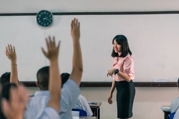 Gülümseyen Asyalı bir bayan lise öğretmeni sınıftaki beyaz üniformalı öğrencilere sorular sorarak ders veriyor ve sonra öğrenciler cevap için ellerini kaldırıyor..