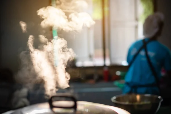 The mass of steam reflected in the morning light coming out of a large electric rice cooker boiling in the cafeteria while the cook is working.