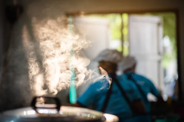 The mass of steam reflected in the morning light coming out of a large electric rice cooker boiling in the cafeteria while the cook is working.