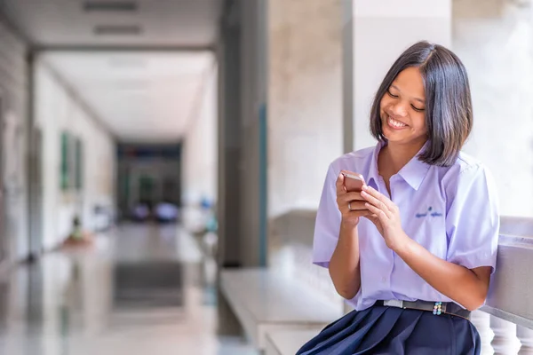 Üniformalı Asyalı kız lise öğrencisi gülümsüyor çünkü akıllı telefonuyla sosyal medyayı sarıyor..