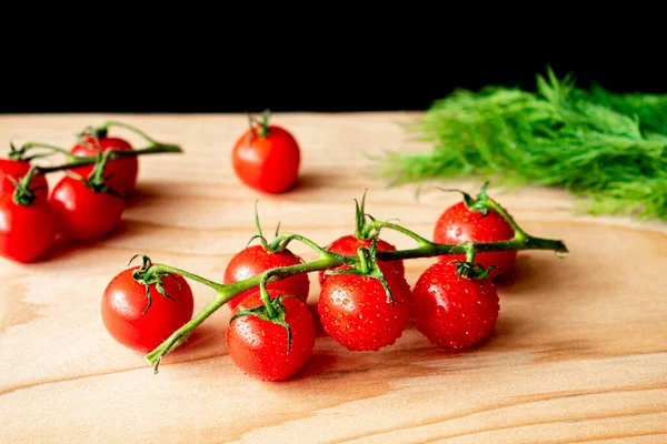 Red cherry tomatoes on a branch on a light wooden Board. In the background there are some more cherry tomatoes and dill