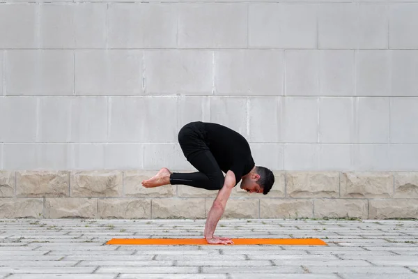 Latin man praktiserar yoga utomhus i en stad, stående i kran pose på orange matta, med grå vägg i bakgrunden. — Stockfoto