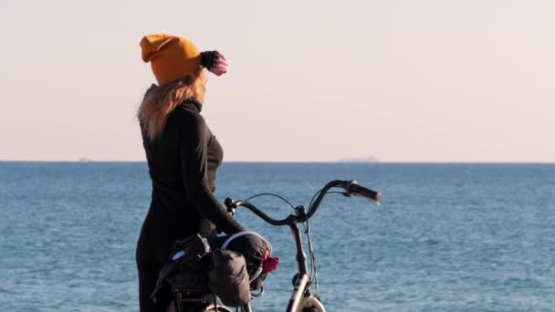 一个头戴黄帽的金发女孩，拿着自行车，望着大海和日落，遮住了阳光的双眼. — 图库视频影像