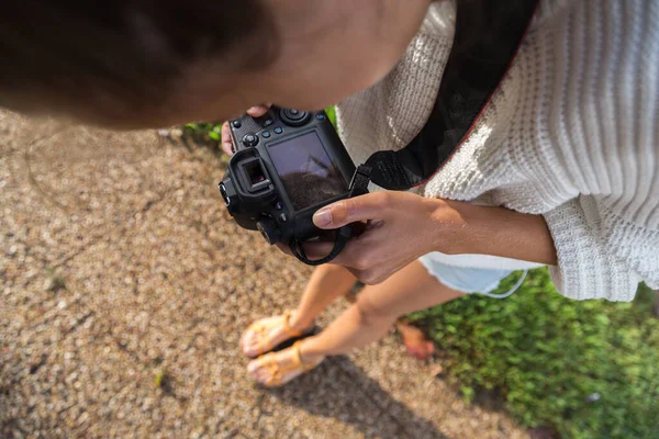 En flicka med reflex kamera tar bilder av hennes ben i sandaler, står på trottoaren med grön gräsmatta — Stockfoto