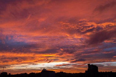 A vibrant sunset with shades of orange, pink, and purple clouds. Silhouettes of buildings are visible in the foreground against the colorful sky.