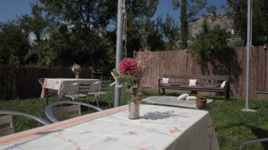 Masası ve sandalyeleri olan güneşli bir bahçe. Masada çiçekli bir vazo var. Yeşil çimenler ve ağaçlar bölgeyi çevreleyerek rahatlatıcı bir atmosfer yaratıyor..