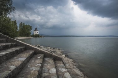 Gölde Liptovska Mara Slovakya ile ön planda taş merdivenle kilise ile kayalık plaj