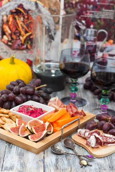 Plate with autumn snacks: beetroot dip , figs, grape, pumpkin, prosciutto, sausage, carrot and two colored glasses of red wine on rustic wooden background.