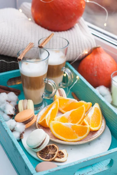 Two cups of latte macchiato on turquoise tray. Plate with grated oranges, macarons, cinnamon rolls and cotton flowers. Knitted sweaters and pumpkins near the window.