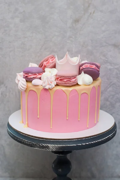 Birthday pink cake for a little girl with fondant crown, melted white chocolate, meringue and cake pops on grey background.