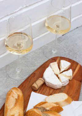 Camembert peyniri, baget ve tahta üzerinde iki bardak beyaz şarap dilimleri. Arkaplan olarak beyaz tuğla duvar.