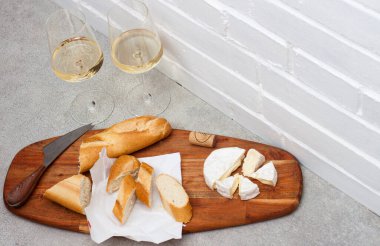 Camembert peyniri, baget ve tahta üzerinde iki bardak beyaz şarap dilimleri. Arkaplan olarak beyaz tuğla duvar.