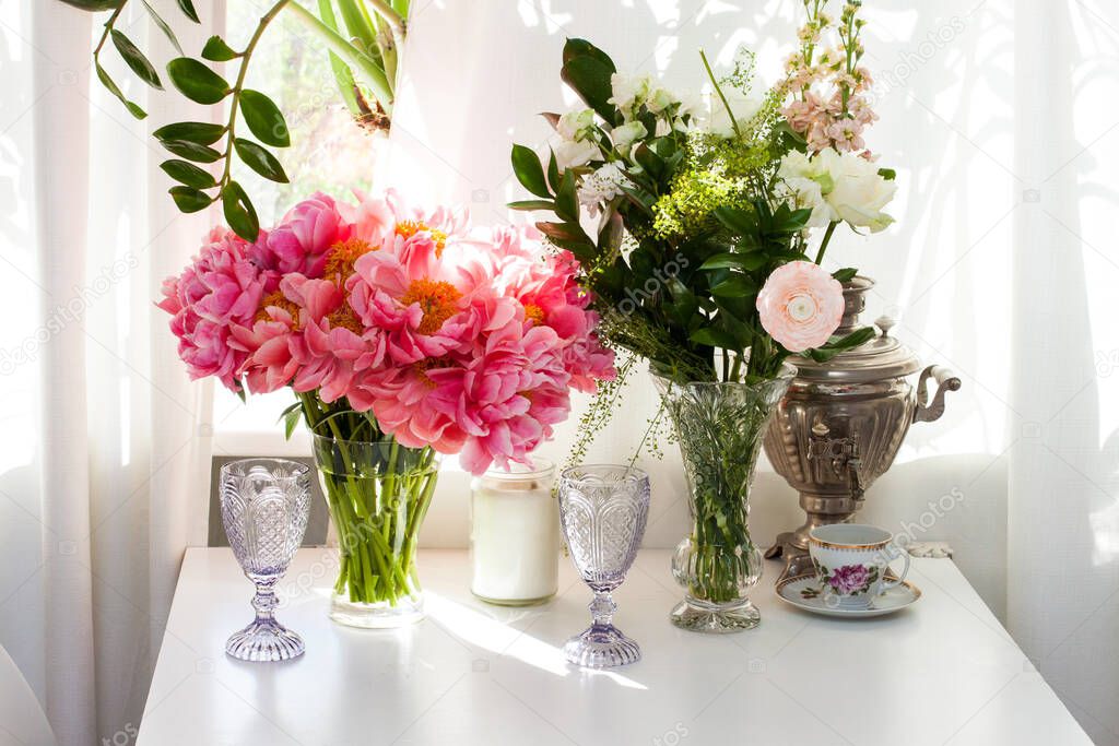 Bouquet of peonies and roses, black and white candles, purple wine glasses and old tea pot on white table near the window.