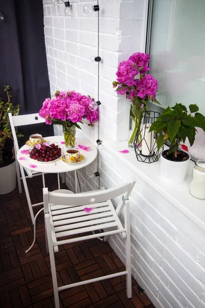 Boeket pioenbloemen, twee kopjes thee en verse kersen op witte tafel. Modern balkoninterieur. Rechtenvrije Stockafbeeldingen