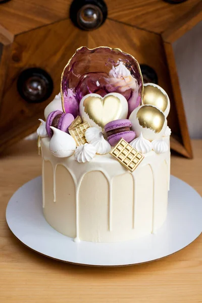 Modern elegant cake with melted white chocolate, golden heart and chocolate balls, macaroons, meringue and caramel.