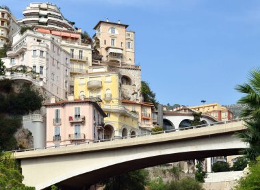 Monte-Carlo, Monaco sokaklarına ve binalara gün ışığında güneşli bir manzara. Park etmiş arabalar ve kaldırım kenarında yürüyen insanlar. Parlak mavi gökyüzü ve arka planda birkaç bulut.