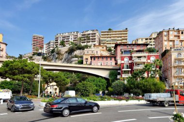 Monte-Carlo, Monaco sokaklarına ve binalara gün ışığında güneşli bir manzara. Park etmiş arabalar ve kaldırım kenarında yürüyen insanlar. Parlak mavi gökyüzü ve arka planda birkaç bulut.
