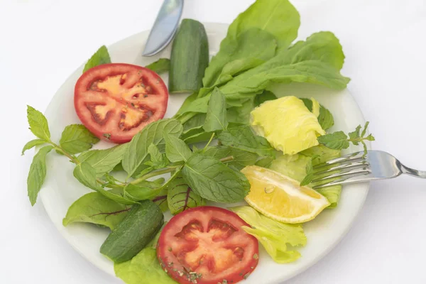 Salading from spinach, mint, mangold, greens, lemon, tomato and cucumber slices on the white plate with knife and fork on white background. Part of low-calorie diet and the key to improvement of digestion