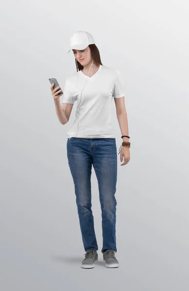ブルーデニムジーンズパンツを着た白い無地VネックTシャツに美しい女性モデルを立っている 帽子をかぶってイヤフォンを手に携帯電話を持っていました隔離された背景 — ストック写真
