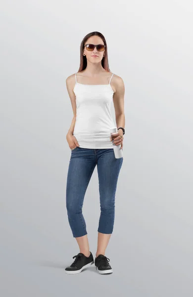 Standing beautiful female model in plain white camisole shirt wearing blue denim jeans pant. Holding white water bottle in hand. Isolated background.