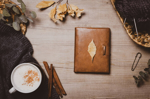 Осенний зимний фон. Старая кожаная тетрадь, чашка капучино, осенние листья, шерстяной шарф и клубок шерстяной пряжи на деревянном фоне. Домашние увлечения, вязание. Уютная концепция комфорта
