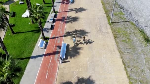 Luchtvlucht achter de jonge man en meisje rijden een fiets langs de promenade — Stockvideo