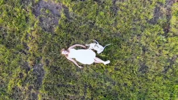 Uma menina bonita jaz na grama com seu cão dálmata. Tomado por drone — Vídeo de Stock