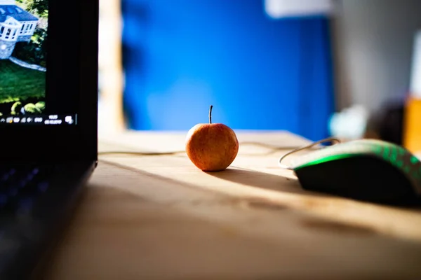 Small red apple on a wooden table with a laptop and a computer mouse