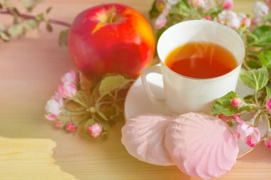 Ahşap bir arka planda, bir dal elma çiçeği, bir kupa çay, marşmelov ve kırmızı elmalar. Durgun hayat. Yakın çekim, yiyecek ve bahar konsepti, kartpostal. Boşluğu kopyala, metin için boşluk.