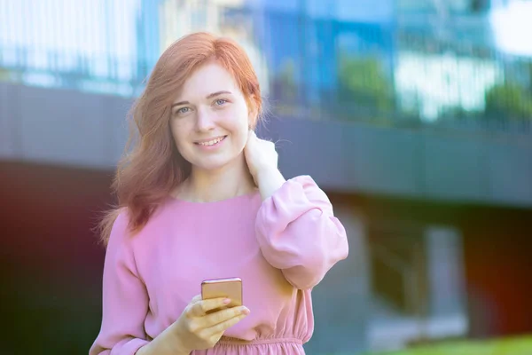 Kızıl saçlı mutlu kadın elinde akıllı telefonuyla kameraya bakıyor ve dışarıda gülümsüyor.