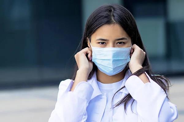 Asian woman, attractive ethnic girl wearing a medical mask on her face, looking at the camera with a serious look in a white shirt outside the business center.