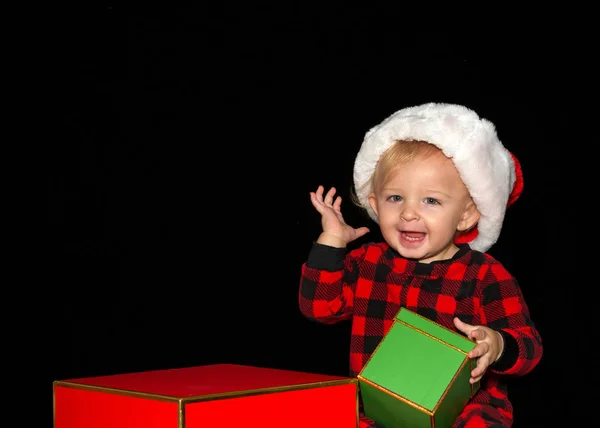 Close up portrait of one one year old baby boy wearing a Santa hat playing with red and green gift boxes, one hand up big smile with teeth showing. Black background with copy space.