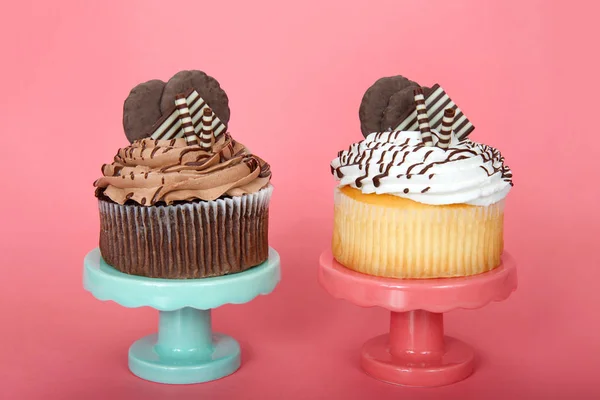 Giant vanilla an chocolate cup cakes with vanilla frosting embellished with white and dark chocolate wafers and cookies on a pink and green pedestals on pink background with copy space