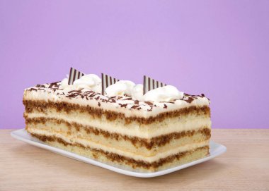 Tiramisu dikdörtgen bir tabakta, ahşap bir masada, mor arka planda. Tiramisu, kahve, brendi ve likörle kaplı katmanlı bir İtalyan tatlısıdır..