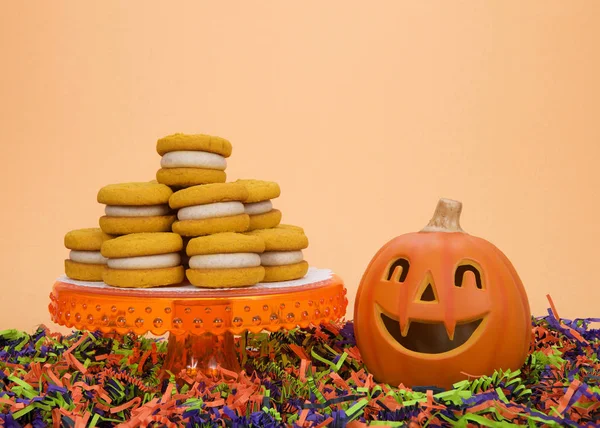 Naturally  flavored Pumpkin Spice cookies with cinnamon cream cheese filling on an orange pedestal with pumpkin jack-o-lantern black orange and green confetti underneath. Orange background copy space