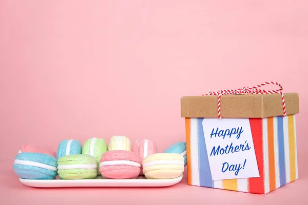 White rectangular plate with pastel colored macaron cookies laying on a pink background, colorful striped present with Happy Mother\'s Day note attached.