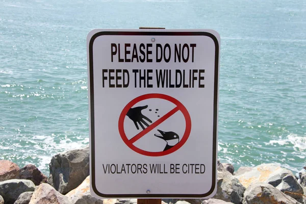 Sign with text DO NOT FEED THE WILDLIFE along the rocky shoreline in a park. Wild animals who depend on people for food can cause injuries, spread disease, become unable to forage for food to survive.