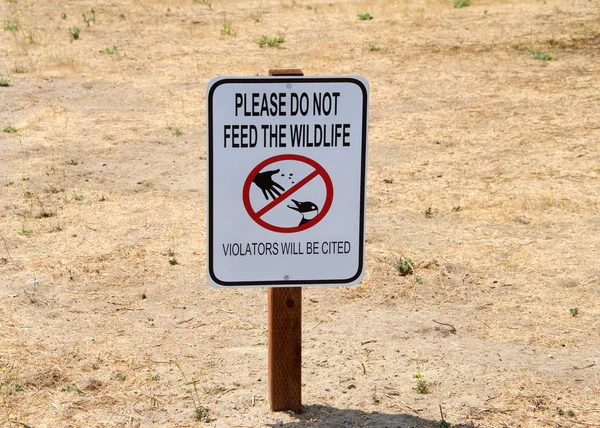 Sign with text DO NOT FEED THE WILDLIFE in a patch of dead grass at a park. Wild animals who depend on people for food can cause injuries, spread disease, become unable to forage for food to survive.