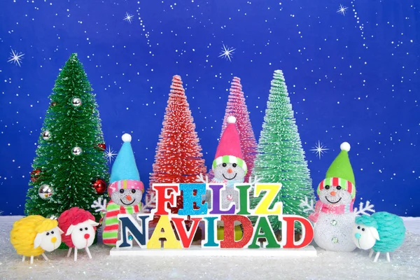 Feliz Navidad, Merry Christmas in Spanish colorful wood sign, red, pink, green pine trees with snowman and colorful yarn sheep on faux snow blue background white dots and stars.